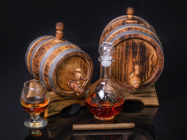 May include: Two wooden barrels with metal bands and spigots, a glass decanter filled with amber liquid, a glass of amber liquid, and a brown cigar on a black surface.
