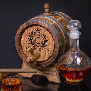 May include: A wooden barrel with a metal band and a spigot, engraved with a crest and the name "C. PERSON". The barrel is on a stand and has a clear glass decanter with a stopper filled with amber liquid.