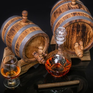 May include: Two small wooden barrels with metal bands and spigots, a glass decanter filled with amber liquid, a glass of amber liquid, and a brown cigar on a black surface.
