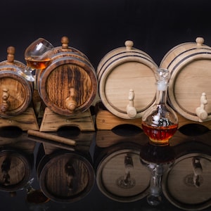 May include: Three wooden barrels with spigots, two larger barrels are side by side on a wooden stand, a smaller barrel is on a separate stand, a glass decanter and a glass with a brown liquid are in front of the barrels.