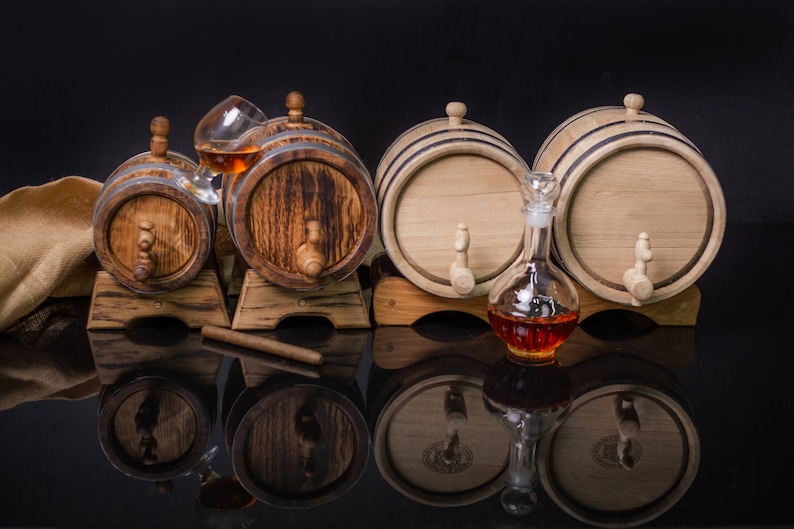 May include: Three wooden barrels with spigots, two larger barrels are side by side on a wooden stand, a smaller barrel is on a separate stand, a glass decanter and a glass with a brown liquid are in front of the barrels.