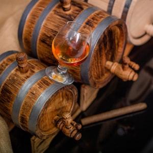 May include: Two wooden barrels with metal bands and spigots, one with a glass of amber liquid inside. The barrels are on a burlap surface.