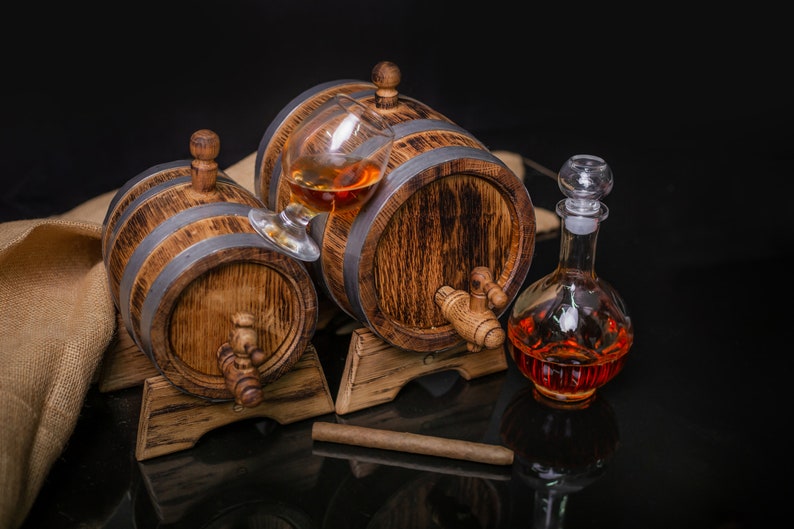 May include: Two wooden barrels with metal bands, one with a glass of amber liquid inside, and a glass decanter filled with amber liquid. The barrels are on wooden stands and there is a brown cigar on the table.