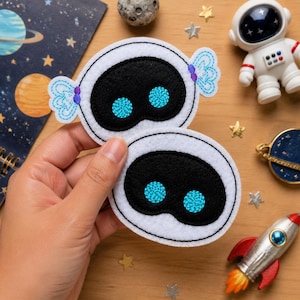 May include: Two white felt patches shaped like robot heads with black oval faceplates and bright blue embroidered circular eyes. The top patch features light blue decorative wing-like accents on the sides with small purple embroidered details.