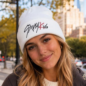 May include: A white knit beanie featuring a folded cuff with the text Stray Kids embroidered in black and red lettering. The word Stray is written in a stylised black script, while the word Kids is written in a simple black font with a red letter K.