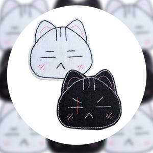 May include: Two cat-shaped felt patches. One is white with black and pink embroidered details, including closed eyes and blush. The other is black with white and pink embroidered details, including a cross and angled lines.