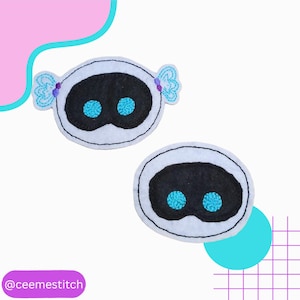 May include: Two white felt appliques with black oval centers and blue embroidered eyes. One applique has blue embroidered details on the sides. The appliques are on a white background with pink and blue geometric shapes. The text "@ceemestitch" is at the bottom.
