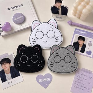 May include: Three felt cat-shaped coasters in white, black, and gray, each featuring embroidered glasses and whiskers. The set is surrounded by Wonwoo Seventeen themed stationery, including a photo card, a notepad, a pen, and a heart-shaped sticker.