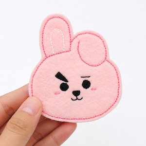 May include: A light pink felt patch shaped like a cartoon rabbit head with one long ear and one folded ear. The face features black embroidered eyes, a small nose, a mouth, and two small pink stitched lines for cheeks, with a pink stitched border.