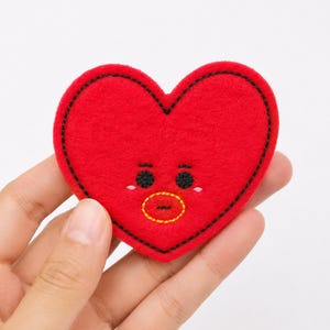May include: A red heart-shaped felt patch featuring embroidered black eyes, a yellow-outlined red mouth, and small white highlights under the eyes. The entire perimeter of the heart is outlined with a thick black stitched border.