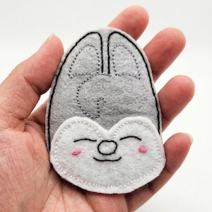 May include: A gray and white felt patch shaped like a stylized animal head with pointed ears. The face features embroidered black lines for closed eyes, a small round nose, a curved mouth, and two small pink stitched circles for rosy cheeks.