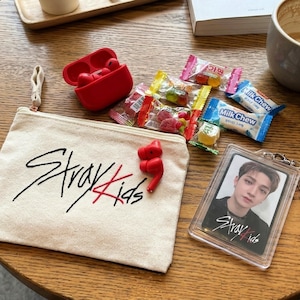 May include: A beige canvas zip pouch with the black and red text Stray Kids, a red wireless earbud case, a pair of red earbuds, several small individually wrapped sweets, and a clear plastic photo card holder featuring a portrait of a person.