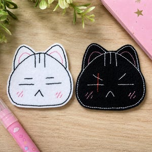 May include: Two embroidered cat face patches, one white and one black, featuring stitched facial expressions with closed eyes, small mouths, and pink blush marks. The black patch includes a red stitched cross mark on the forehead area.