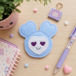 May include: A light blue felt patch shaped like a head with two rounded ears and a white heart-shaped face. The patch features embroidered dark purple heart-shaped eyes and a small black smile, with light pink stitched blush marks on the cheeks.