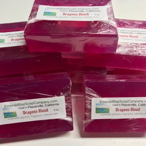 Op de afbeelding: Verschillende blokken Dragons Blood zeep, afzonderlijk verpakt in doorzichtig plastic. Elk blok heeft een etiket met de tekst "EmeraldBaySoapCompany.com" en "Dragons Blood". De zeep bevat plantaardige glycerine, geur en natuurlijke kleur.