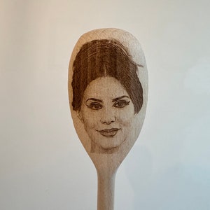 Puede incluir: Cuchara de cocina de madera con un retrato grabado de un rostro femenino. El retrato está centrado en la cabeza ovalada de la cuchara, mostrando los rasgos de la mujer. La cuchara está hecha de madera clara.