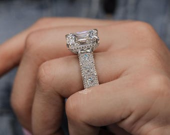 May include: A close-up of a silver ring with a large, rectangular, clear gemstone. The ring has a detailed, ornate design with smaller gemstones set into the band and around the main stone. The ring is on a finger.