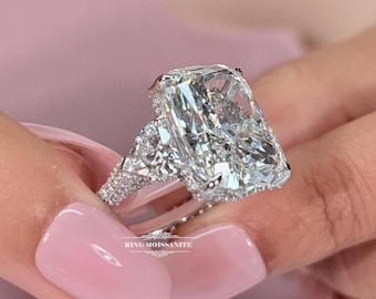 May include: A radiant-cut engagement ring with a large, rectangular center stone and two triangular side stones. The ring is set in a silver band and is held up by a person's fingers. The text "RING MOISSANITE" is visible.