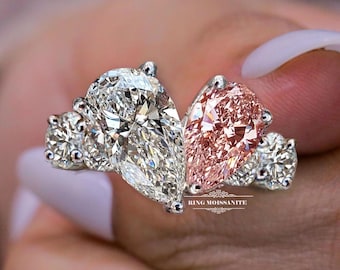 May include: A close-up of a ring featuring a large, clear pear-shaped stone and a smaller, pink pear-shaped stone. The ring has a silver band and smaller round stones on either side of the larger stones. The text "RING MOISSANITE" is visible.
