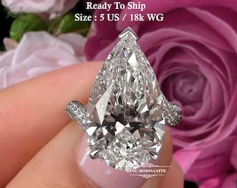 May include: A pear-shaped diamond ring with a brilliant cut. The ring is set in white gold and features smaller accent stones on the band. The image includes the text "Ready To Ship" and "Ring Moissanite".