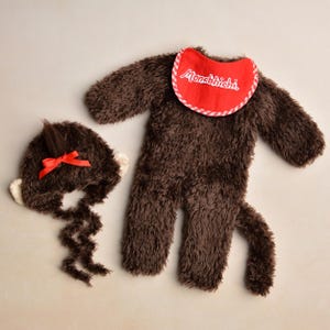 Könnte beinhalten: Ein braunes Plüsch-Affenkostüm-Set für Babys, einschließlich eines Overalls, eines Hutes mit roter Schleife und einer Lätzchen mit dem Text "Monchhichi". Das Kostüm hat einen Schwanz und Ohren.