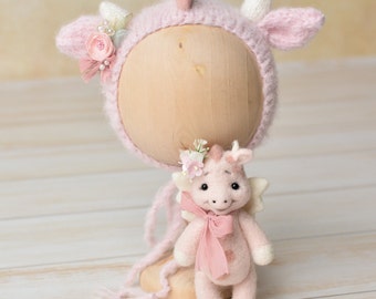 May include: A pink knitted hat with a unicorn horn and ears, decorated with flowers and pearls. A pink felt unicorn toy with wings and a pink bow is sitting on a wooden stand.