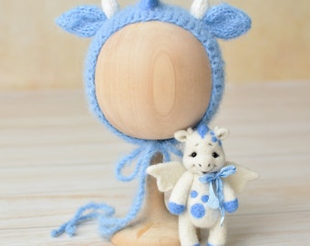 May include: A light blue knitted baby bonnet with white horns and ear accents, displayed on a wooden head form. A small white and blue dragon plush toy with wings and a blue bow is next to the bonnet.