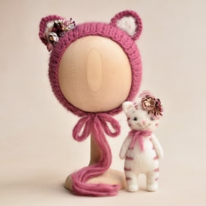 May include: A knitted magenta baby bonnet with cat ears and floral accents, displayed on a wooden head form. A matching white and magenta cat doll with a floral headpiece and pink scarf is next to the bonnet.