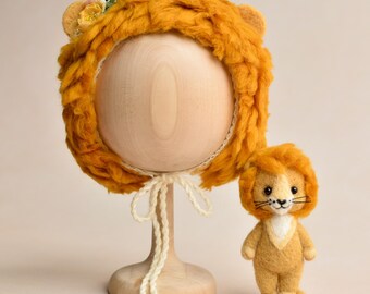 Op de afbeelding: Een pluche, goudgele leeuwenhoed met aangehechte oren en een bijpassende knuffel leeuw. De hoed wordt op een houten standaard getoond en heeft een crèmekleurige stropdas. De knuffel leeuw heeft een witte borst en een zwart gestikte neus en snorharen.