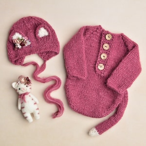 May include: A knitted baby set in a deep pink color, including a hat with ear details and floral embellishments, a long-sleeved onesie with wooden buttons, and a small stuffed cat toy. The onesie has a tail with a white tip.