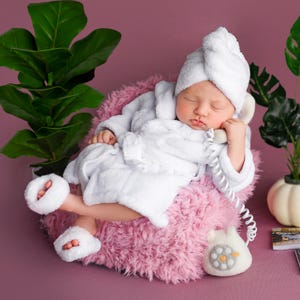 May include: A newborn baby wearing a white terry cloth robe, towel turban, and slippers, resting on a fluffy pink cushion. The baby is holding a white telephone receiver to its ear. A felt telephone and plants are nearby.