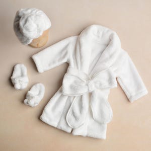 Può includere: Una vestaglia di peluche bianca con un fiocco, una fascia per capelli in peluche bianca e due pantofole di peluche bianche.