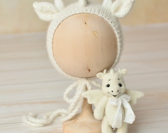 May include: A white knitted unicorn hat with a long white tie, displayed on a wooden stand. A small white knitted dragon with wings and a white satin ribbon is standing next to the hat.