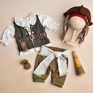 Puede incluir: Un conjunto de disfraz de pirata con una camisa blanca con volantes, un chaleco a cuadros con detalles de cuero marrón, pantalones verde oliva con una faja a rayas, un sombrero marrón con trenzas y un catalejo de juguete. También se incluye una brújula.