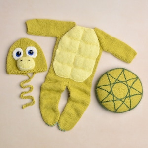 Op de afbeelding: Een gebreide baby-outfitset in tinten geel en groen, met een schildpadontwerp. De set bevat een hoed met een groot blauwogig gezicht, een eendelig pak met een schildpadschildontwerp en een rond kussen met een sterrenpatroon.