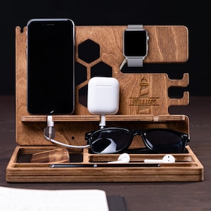 Può includere: Una stazione di ricarica in legno con più scomparti. Contiene uno smartphone, uno smartwatch, auricolari wireless e occhiali da sole. La stazione ha una finitura in legno scuro e include un piccolo blocco note e una penna. Il nome "WILLIAM" è inciso.