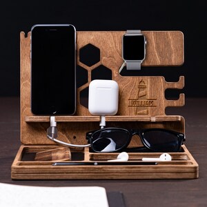 Puede incluir: Organizador de escritorio de madera con compartimentos para smartphone, smartwatch y accesorios. Cuenta con una bandeja para gafas, auriculares y un bolígrafo. El organizador es de madera teñida e incluye el nombre "WILLIAM" grabado en el frente.