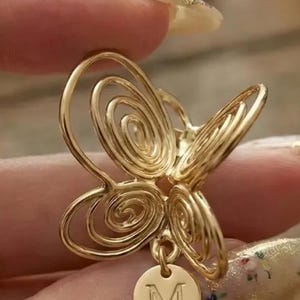 Könnte beinhalten: Goldfarbener schmetterlingsförmiger Anhänger mit Spiraldesign. Ein kleiner, runder Anhänger mit dem Buchstaben "M" hängt unten. Der Anhänger wird zwischen den Fingern gehalten.