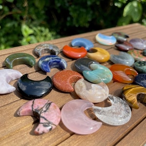 Può includere: Una collezione di 20 pietre levigate a forma di mezzaluna di vari colori, tra cui verde, blu, rosso, nero, bianco, rosa e marrone. Le pietre sono disposte su una superficie di legno.