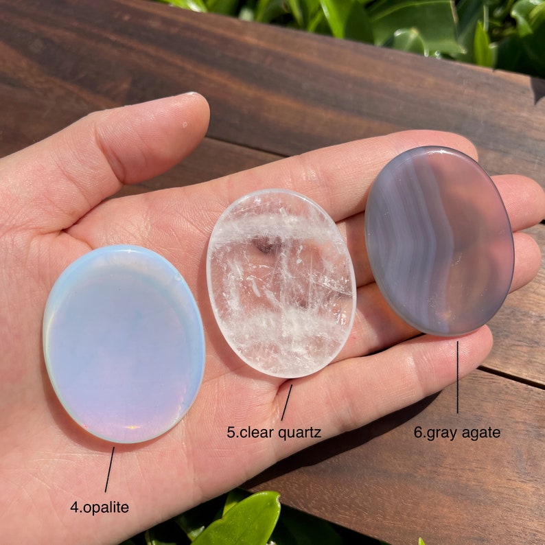 May include: Three oval-shaped polished stones, a clear quartz, a gray agate, and an opalescent stone, are displayed on a hand. The stones are labeled with their names.