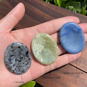 May include: Three smooth, polished palm stones in different colors: a dark gray larvikite, a light green Chinese jade, and a light blue blue aventurine. The stones are arranged in a row on a hand.
