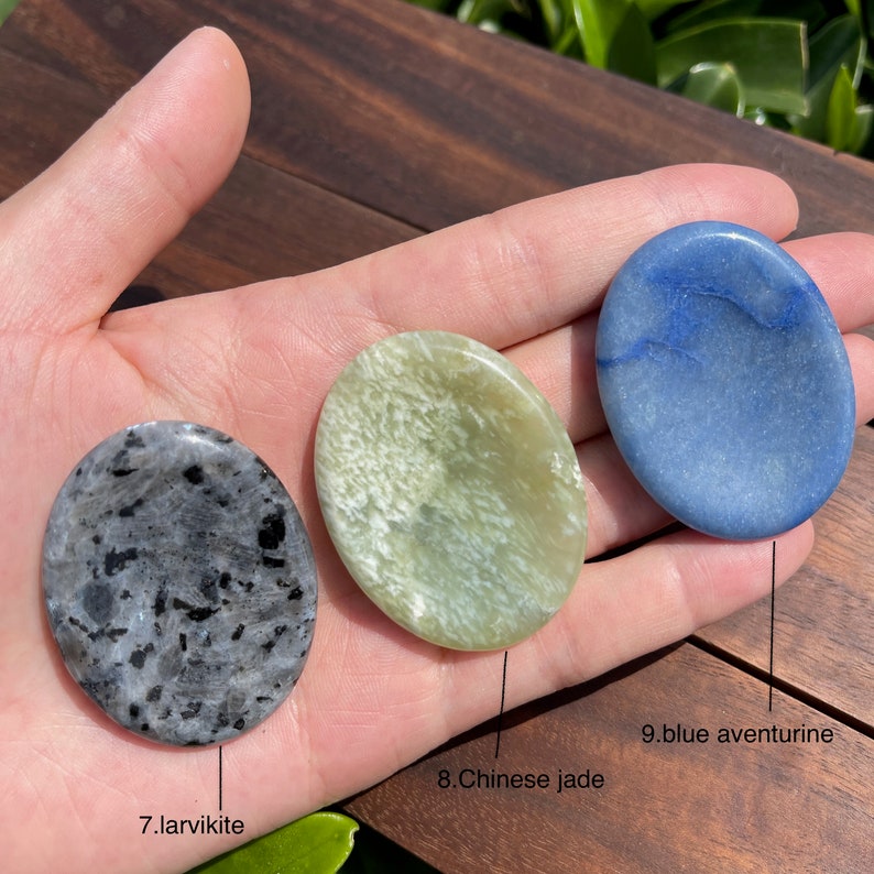 May include: Three smooth, polished palm stones in different colors: a dark gray larvikite, a light green Chinese jade, and a light blue blue aventurine. The stones are arranged in a row on a hand.