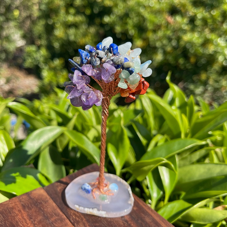 May include: A wire-wrapped crystal tree with a white agate base. The tree is decorated with amethyst, red jasper, green aventurine, blue lapis lazuli, and clear quartz crystals.