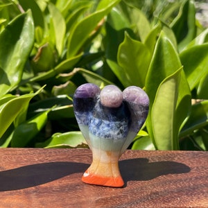May include: A multi-colored angel figurine carved from seven chakra stones. The angel is standing on a wooden surface. The stones are amethyst, sodalite, green aventurine, citrine, carnelian, and red jasper.