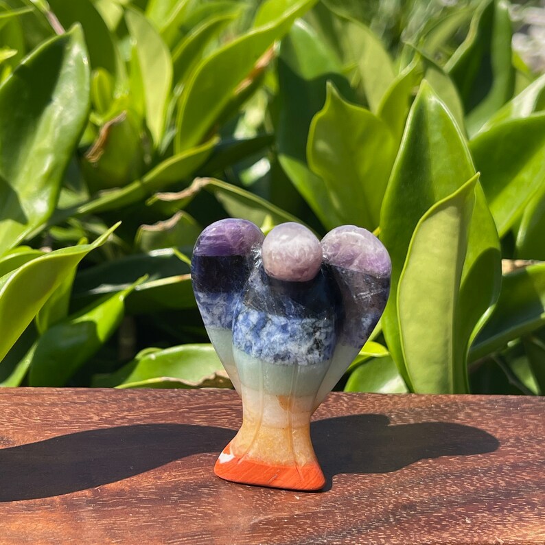 May include: A multi-colored angel figurine carved from seven chakra stones. The angel is standing on a wooden surface. The stones are amethyst, sodalite, green aventurine, citrine, carnelian, and red jasper.