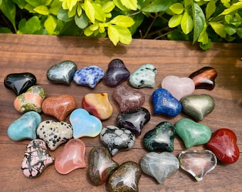 May include: A collection of 20 heart-shaped polished stones in various colors and textures, including green, blue, red, pink, black, and white. The stones are arranged on a wooden surface.