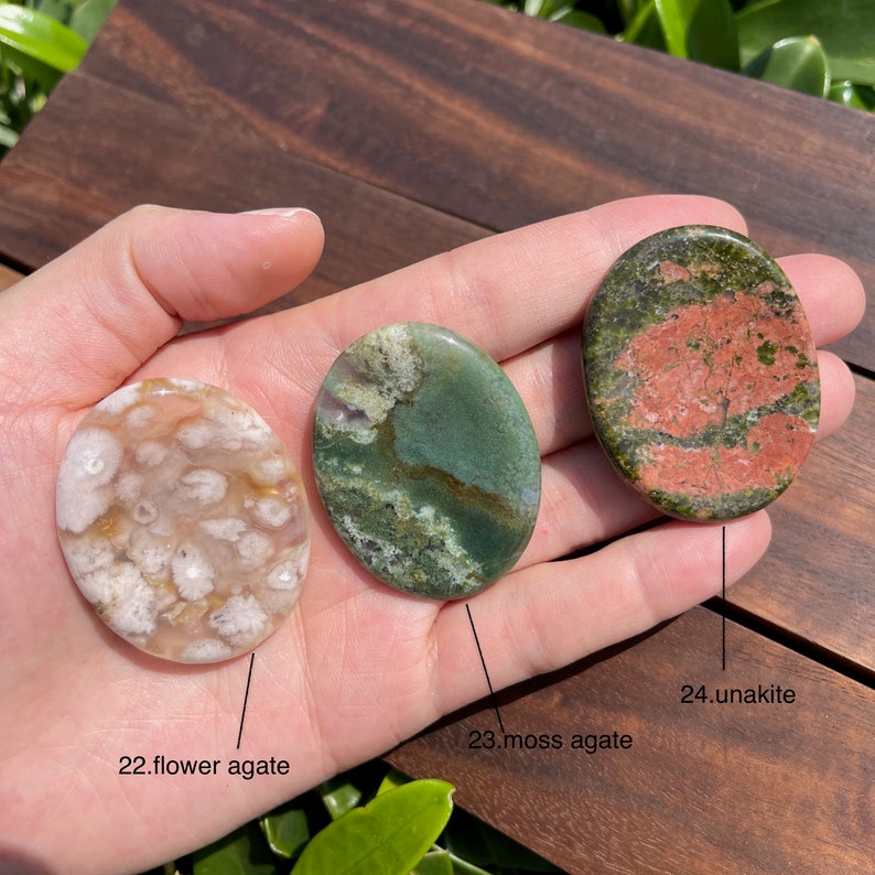 May include: Three polished oval-shaped stones, a pink and white flower agate, a green moss agate, and a red and green unakite. The stones are arranged in a row on a hand.