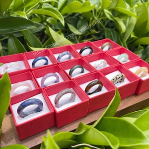 May include: A collection of gemstone rings displayed in small red boxes. The rings are made of various types of stones, including amethyst, rose quartz, and jasper. The boxes are arranged in a grid pattern on a wooden surface. The image includes the text "https://onboardcrystals.etsy.com".