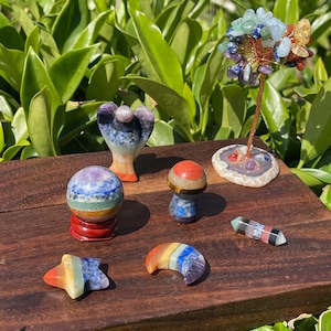 Peut inclure: Sept figurines en cristal de chakra, dont une sphère, un ange, un champignon, une lune croissante, une étoile et une baguette. Les cristaux sont disposés sur une surface en bois.