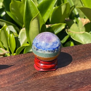 May include: A seven chakra crystal ball on a red wooden stand. The ball is made of layered gemstones in shades of purple, blue, green, brown, and orange.